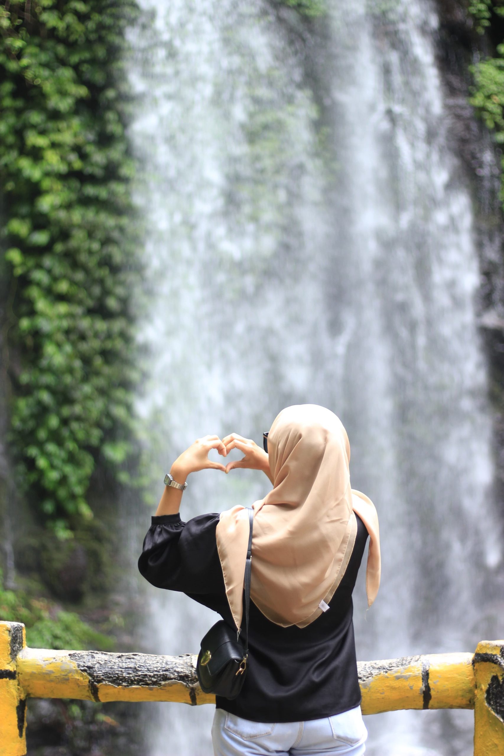 A woman in a hijab forms a heart shape with her hands while admiring a waterfall in Wonogiri, Indonesia.
